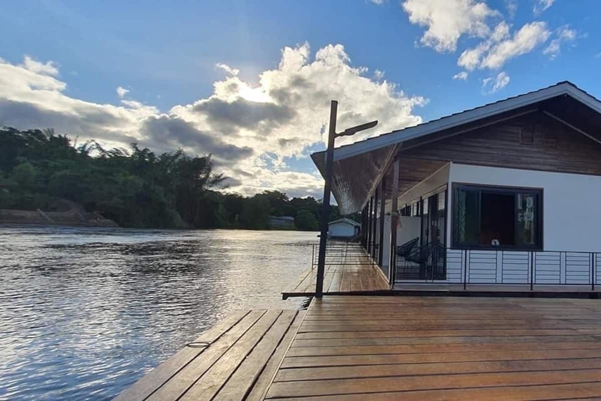 A wooden dock extending into a calm river, leading to a modern house with a sloped roof and large windows.