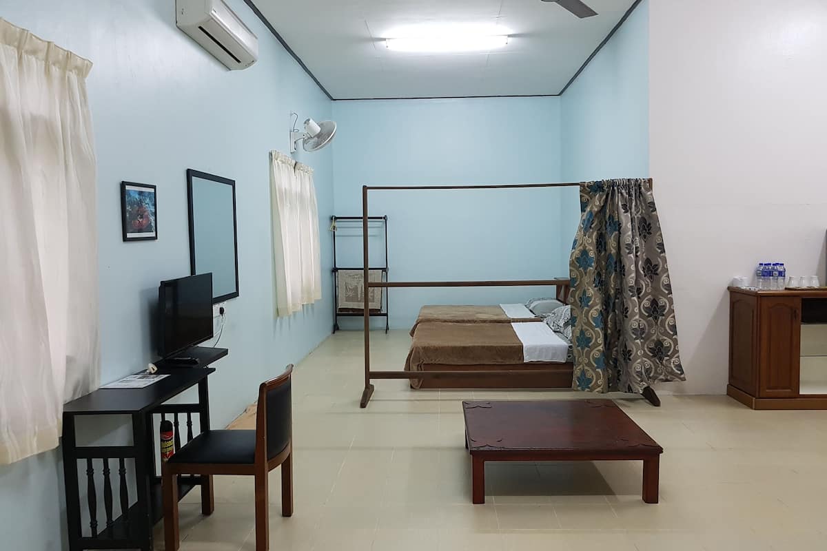 Ein Zimmer mit einem Bett, einem Schreibtisch, einem Sessel, einem kleinen Tisch, einer Klimaanlage, einem Deckenventilator und einem Fenster mit Vorhängen.