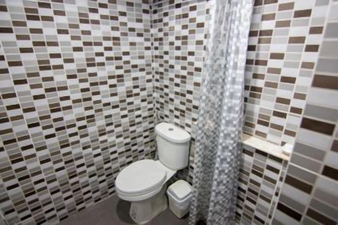 A bathroom with a tiled floor and walls, a toilet, a trash bin, and a shower curtain.
