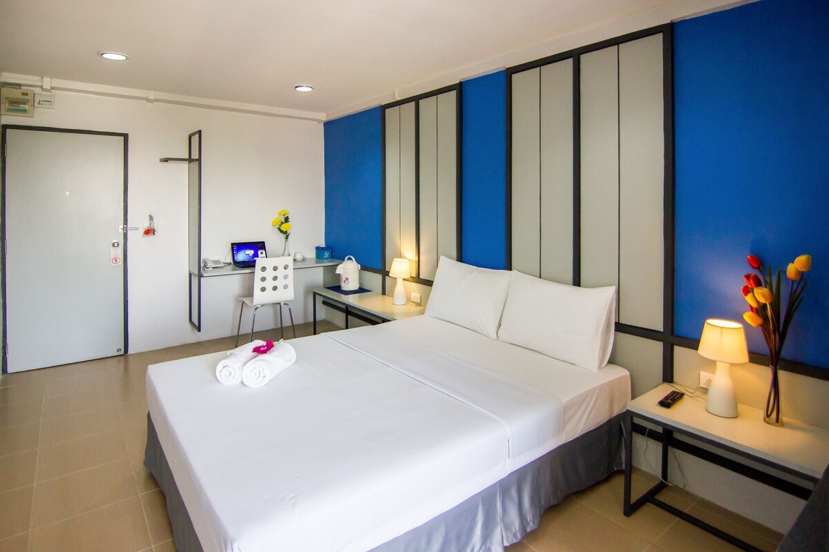 A hotel room with a bed, a desk with a computer, a chair, a nightstand with a lamp, and a wall with vertical panels.