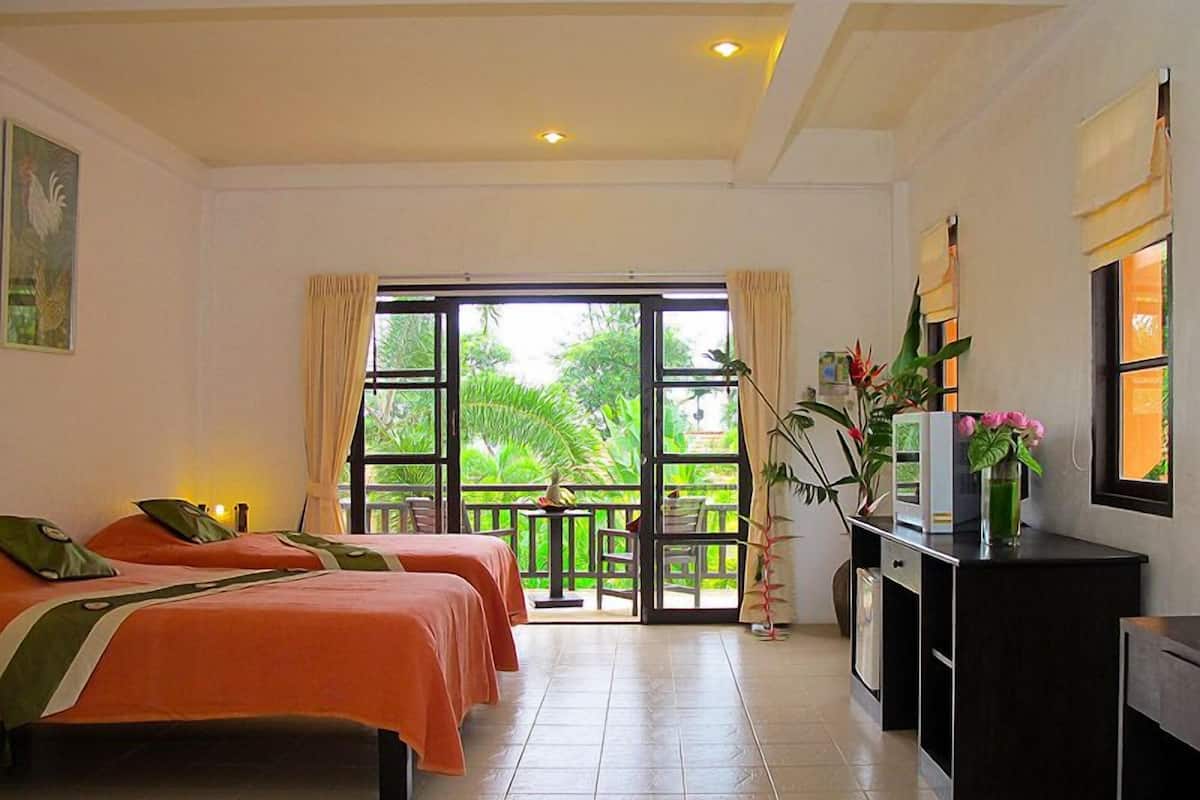 A hotel room with two beds, a desk, a chair, and a balcony with a view of greenery.