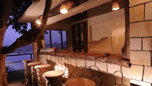 A stone bar with wooden stools, a stone fireplace, and a view of the outdoors. - Pugdundee Safaris - Ken River Lodge (Panna)