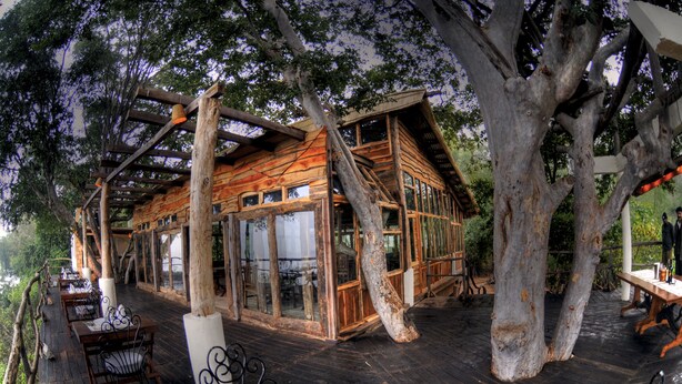 A rustic wooden cabin with large windows and a deck surrounded by trees. - Pugdundee Safaris - Ken River Lodge (Panna)