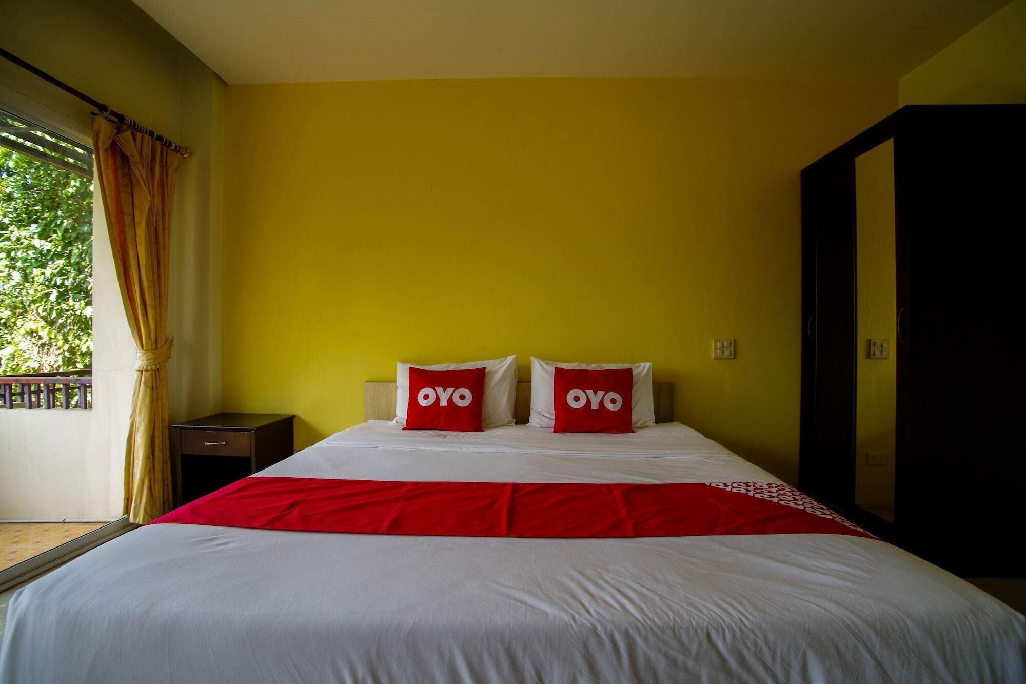 A hotel room with a bed, two pillows with 'OYO' branding, a red and white bedspread, a nightstand, and a wardrobe.
