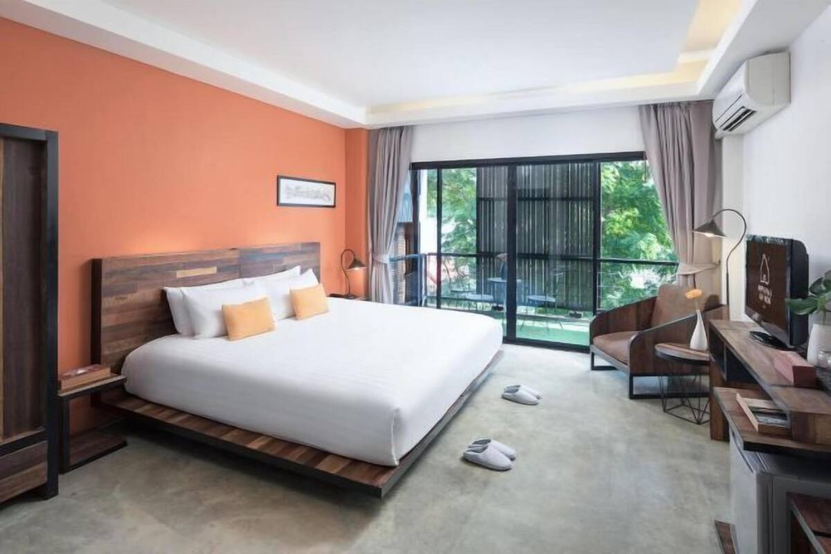 A hotel room with a large bed, a desk, a chair, a TV, and a balcony with a view of greenery.