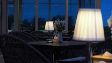 A table with a lamp and a vase with flowers, set against a backdrop of large windows.