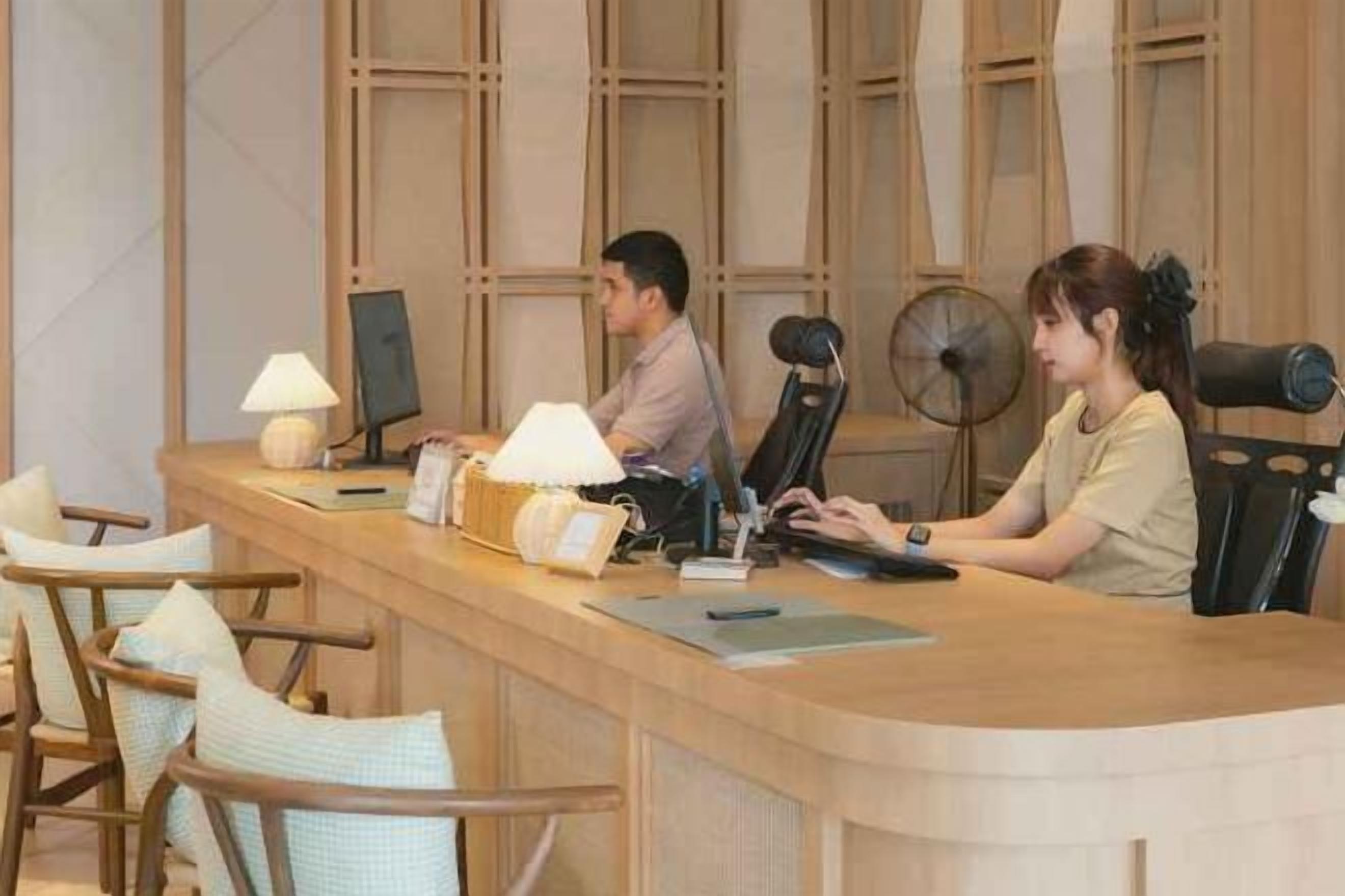 A reception desk with two staff members, a fan, and a lamp.