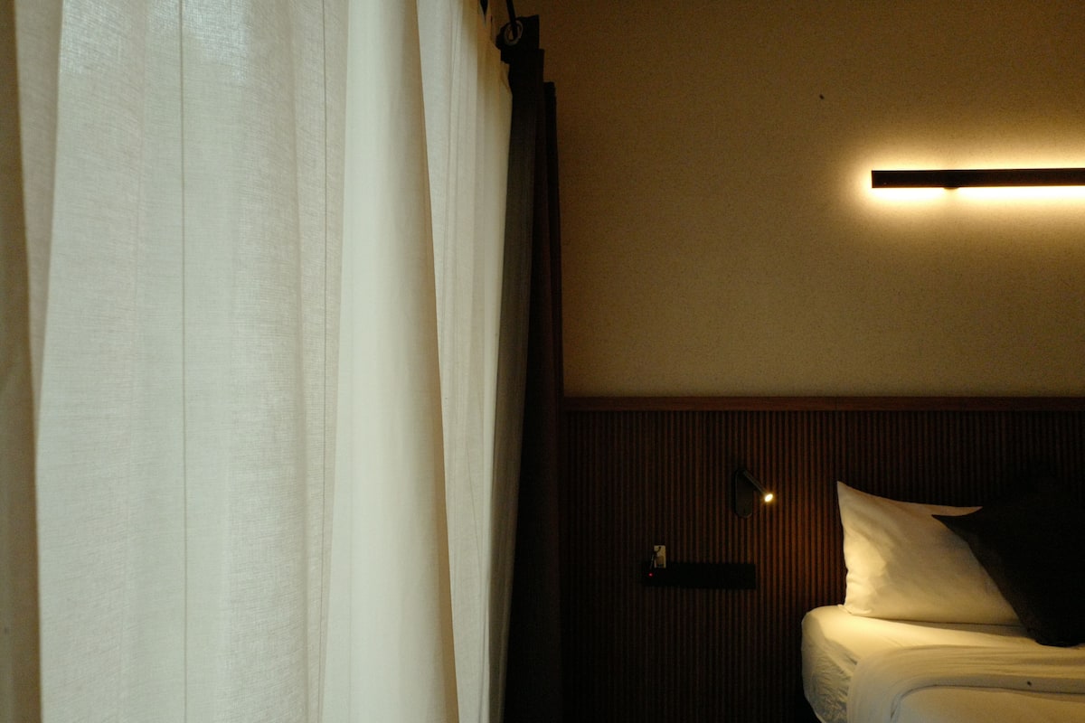 A hotel room with a bed, a nightstand, a wall-mounted light, and a curtain.