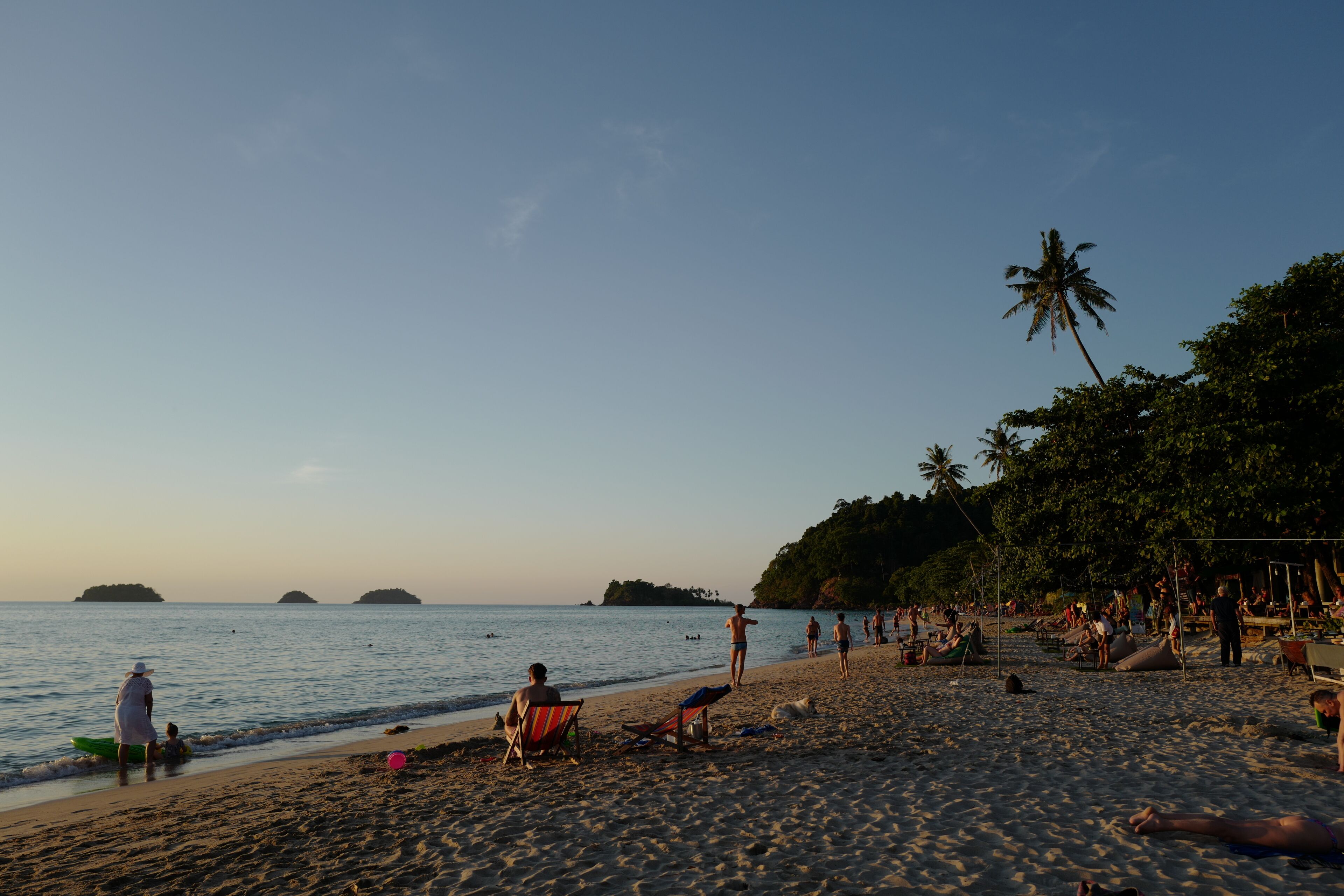 A beach with people relaxing, a few boats, and palm trees.