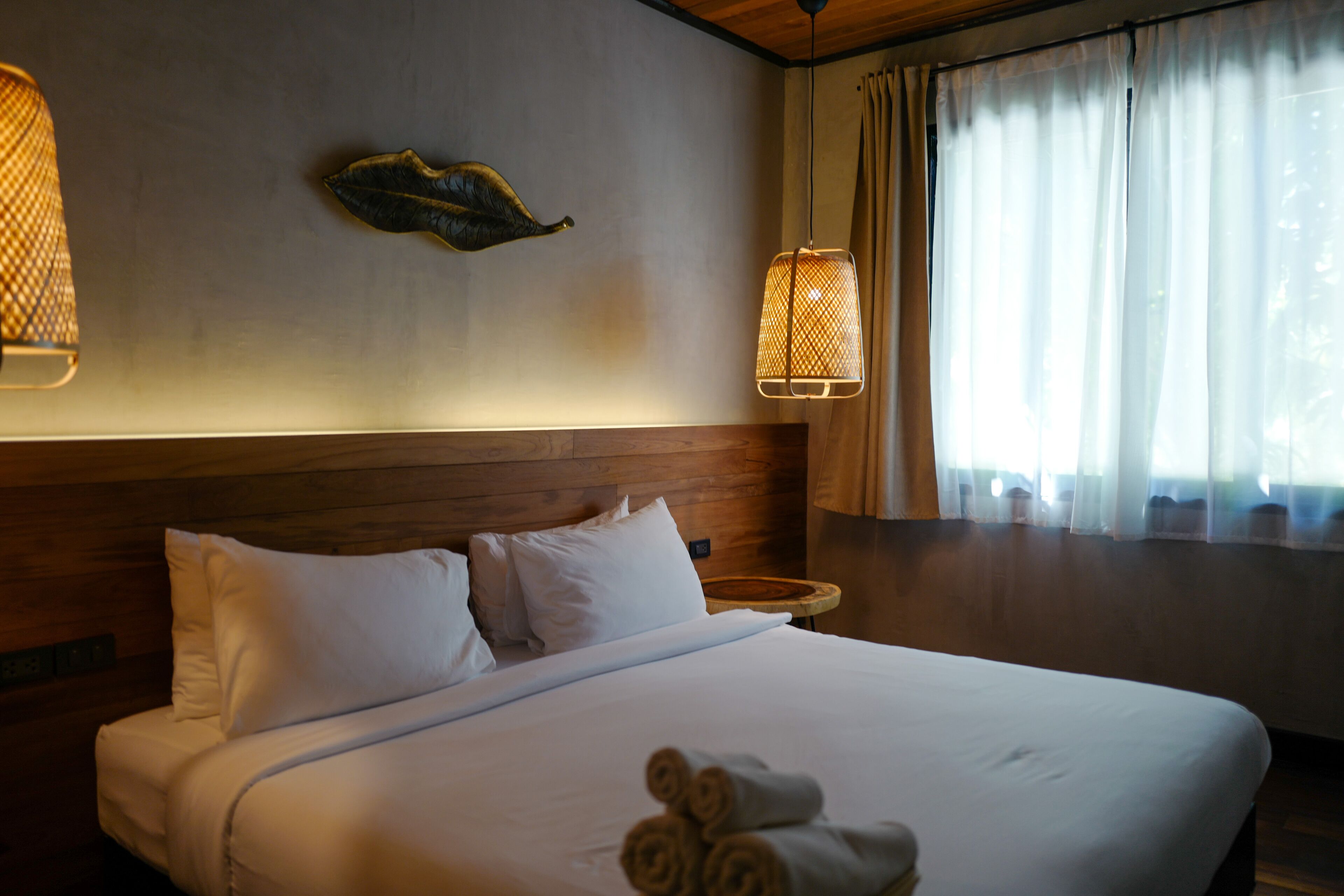 A neatly made bed with white linens and rolled towels, a wooden headboard, and a decorative leaf on the wall.