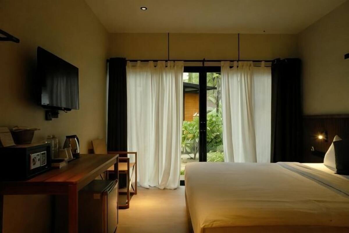 A bedroom with a bed, a desk, a television, and a view of greenery through the window.
