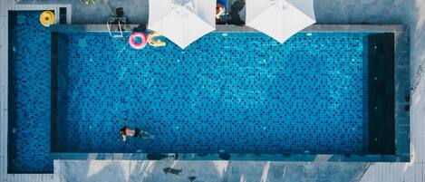 An aerial view of a rectangular swimming pool with tiled sides, surrounded by lounge chairs and umbrellas.