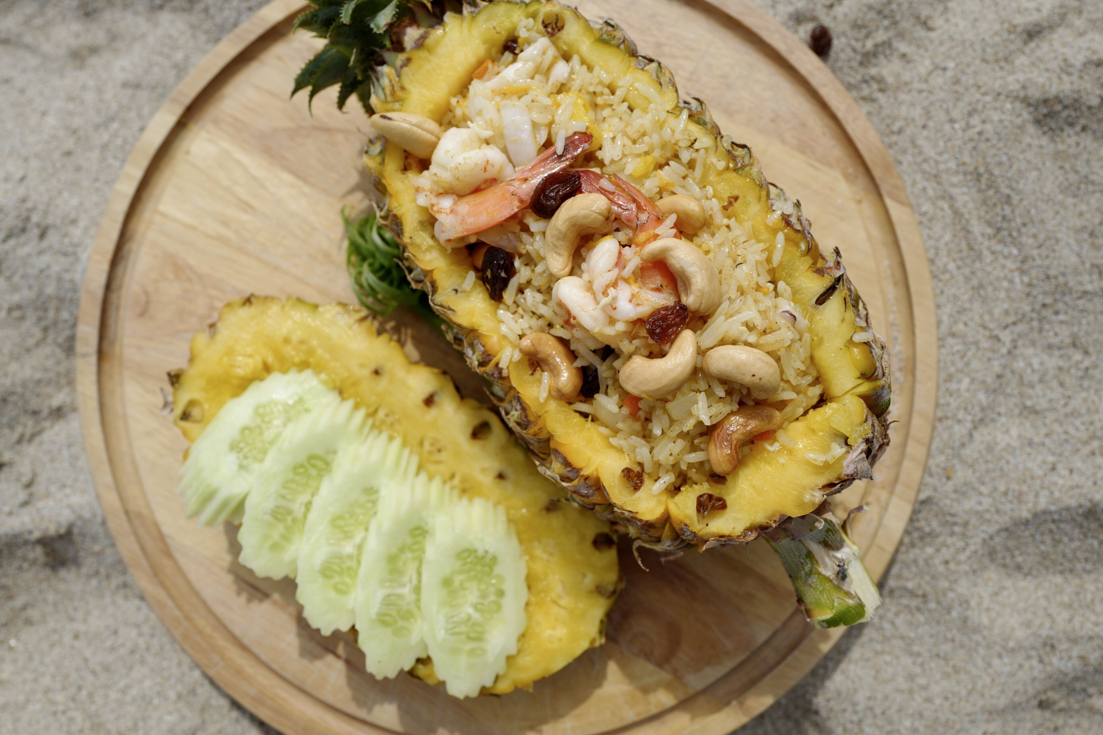 Un plat de fusta amb una meitat d'ananas farcit d'arròs, gambes, cacauets, raïms i rodanxes de pepino.