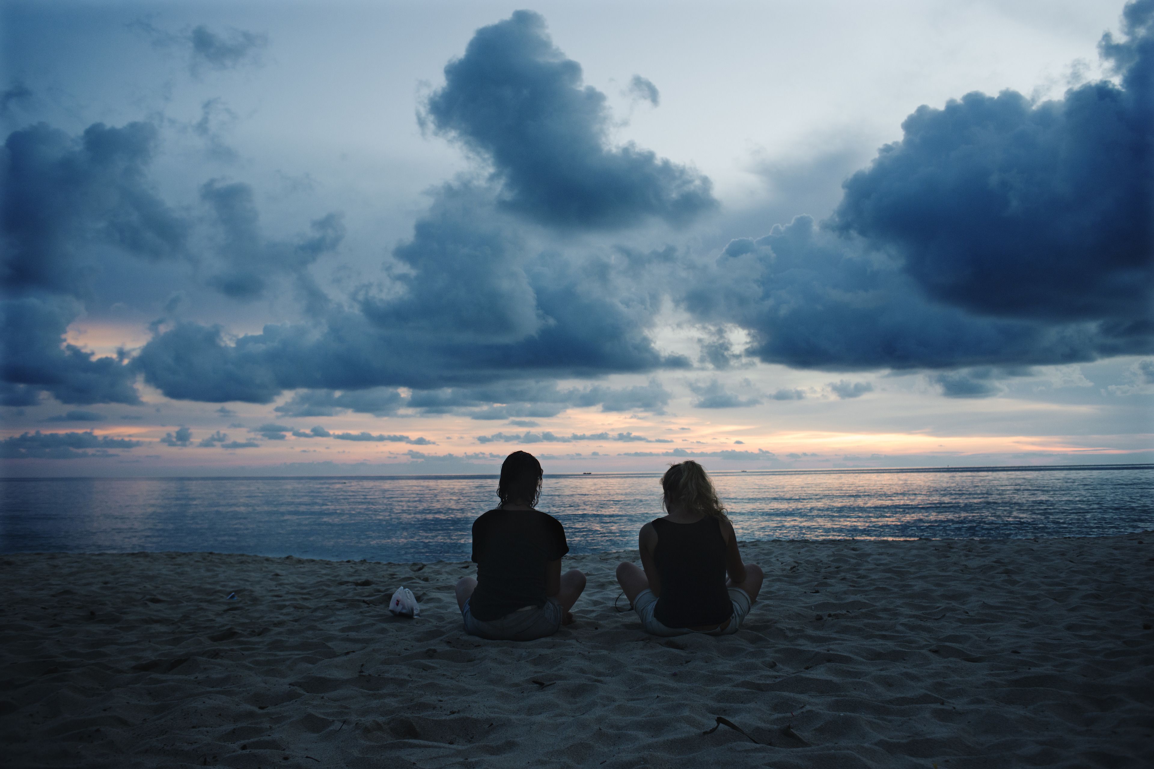 Twee personen zitten op een zanderig strand, uitkijkend over de oceaan onder een bewolkte lucht.