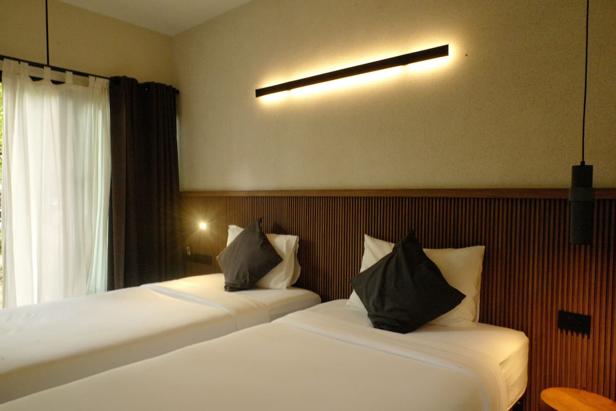 A hotel room with two beds, a wooden headboard, a wall-mounted light, and a window with curtains.
