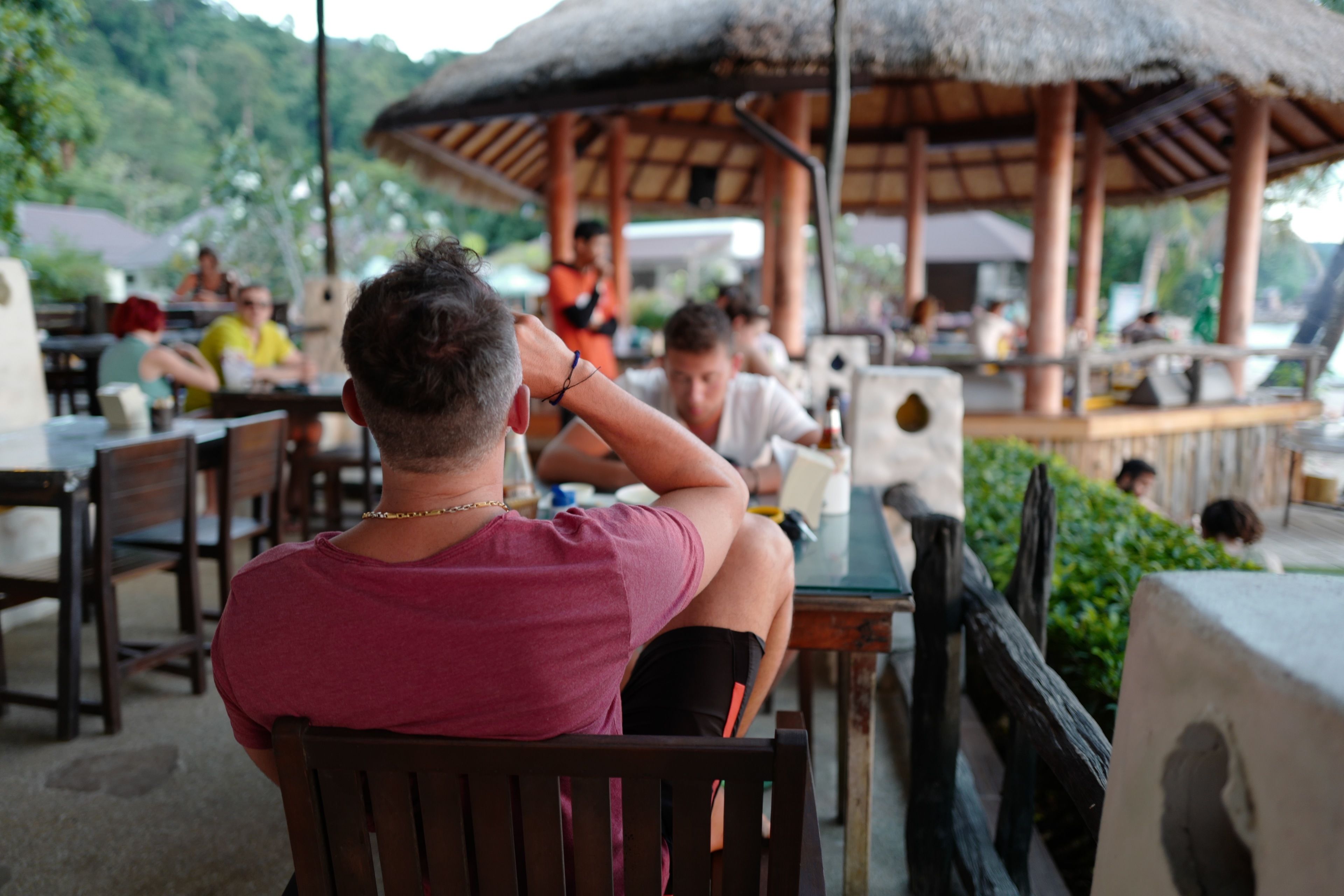 A dining area with wooden tables and chairs, a thatched roof structure, and greenery in the background.