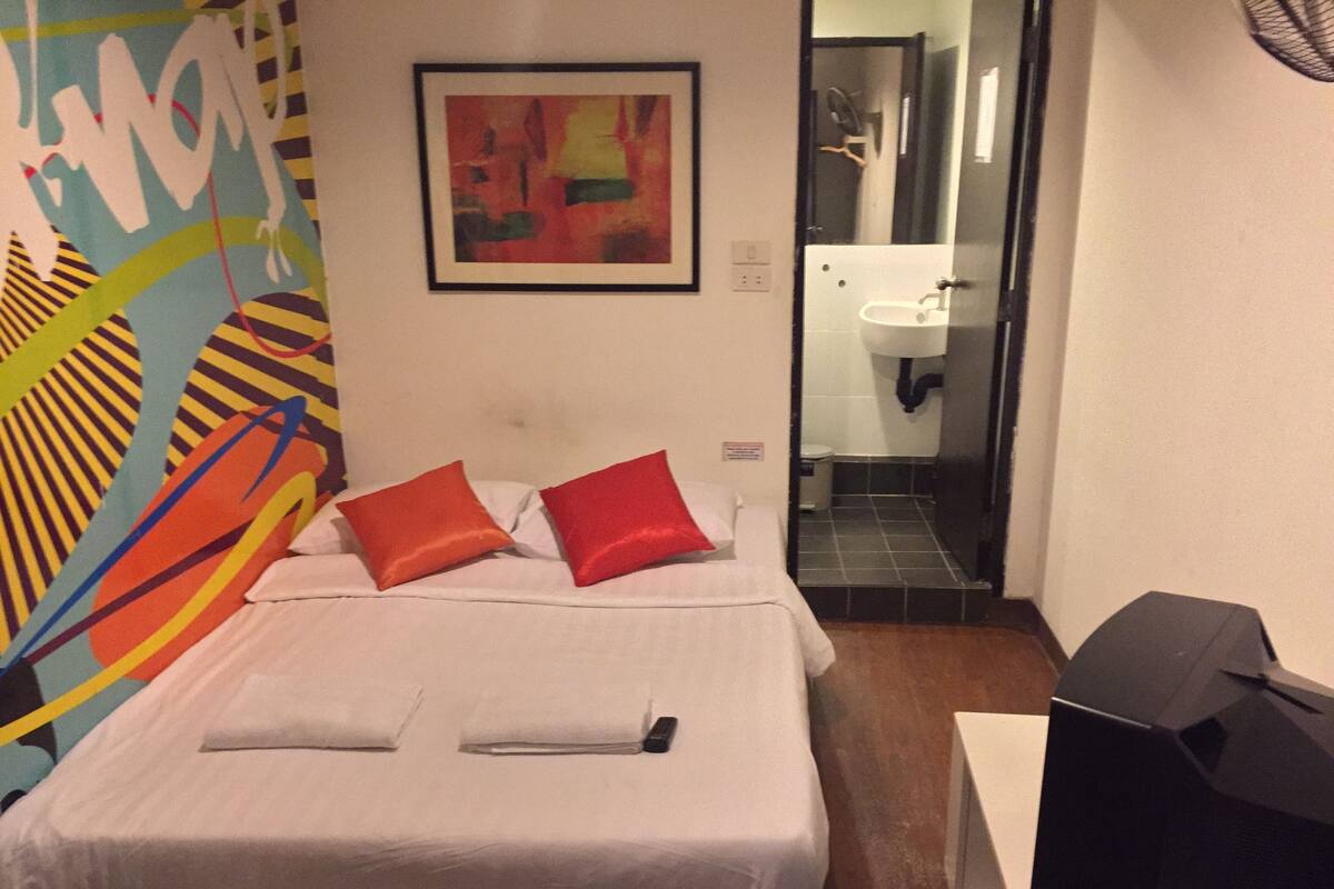 Una habitación de hotel con cama, almohadas, un cuadro, un baño con lavamanos y televisión.