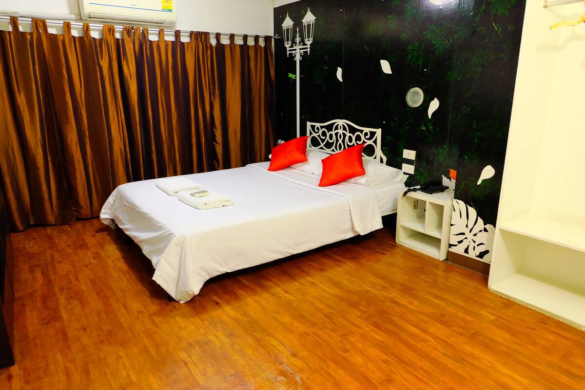 Una cama bien hecha con ropa blanca y dos almohadas rojas, piso de madera, una mesita de noche blanca y una pared con un panel decorativo negro.