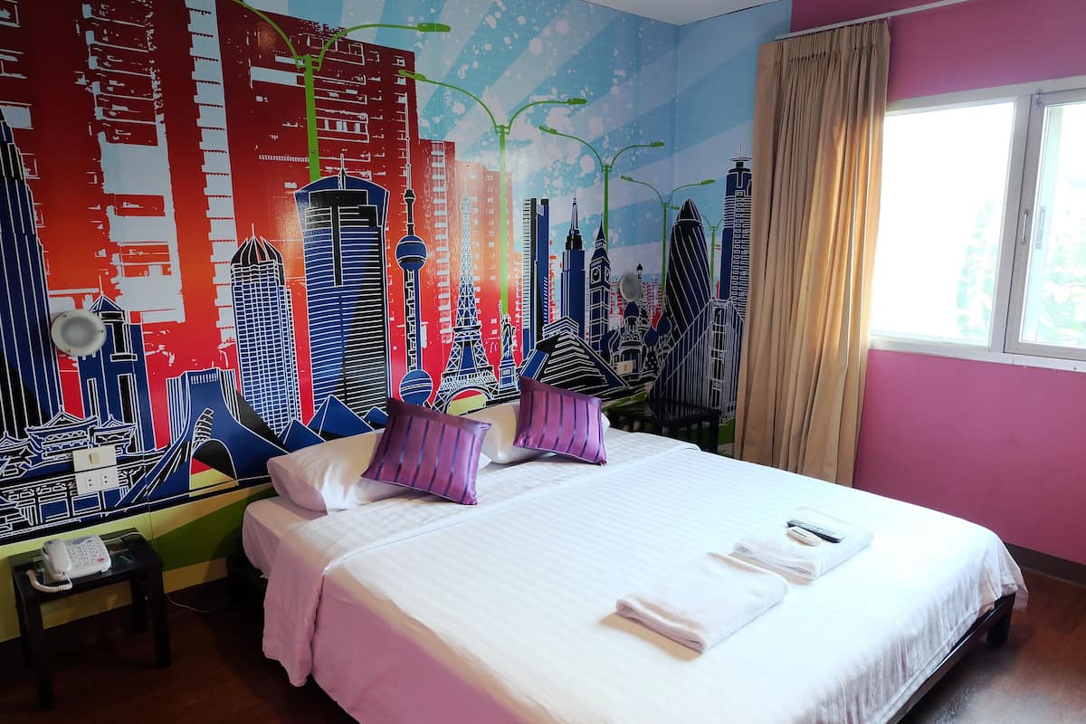 Habitación de hotel con una cama, almohadas, una mesita de noche con teléfono y un mural de un paisaje urbano colorido en la pared.