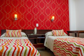 Une chambre d’hôtel avec deux lits, chacun recouvert d’une couvre-lit à rayures colorées. On y trouve deux lampes fixées au mur et une table de chevet avec un miroir et un récipient en verre.