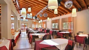 A spacious dining area with multiple tables, each covered with a white tablecloth and red table runners. The room features wooden ceiling beams and hanging pendant lights. - Harmonie Hotel de Montaña (Villa General Belgrano)