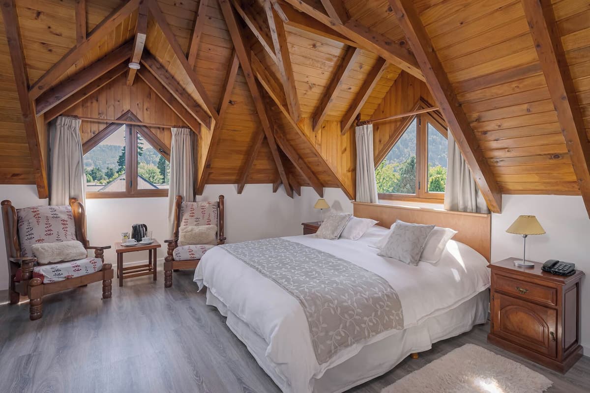 Een gezellige zolderkamer met een groot bed, twee stoelen, een nachtkastje en een raam met gordijnen.