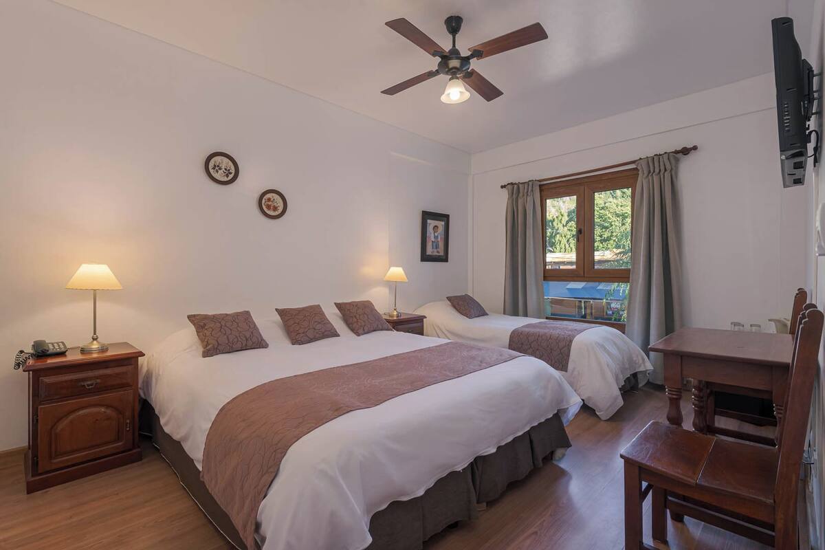 Een hotelkamer met twee bedden, een plafondventilator, een bureau en een raam met gordijnen.