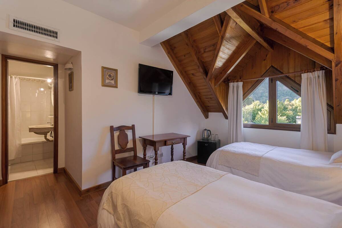 Een slaapkamer met twee bedden, een houten plafond, een televisie, een tafeltje en een raam met gordijnen.