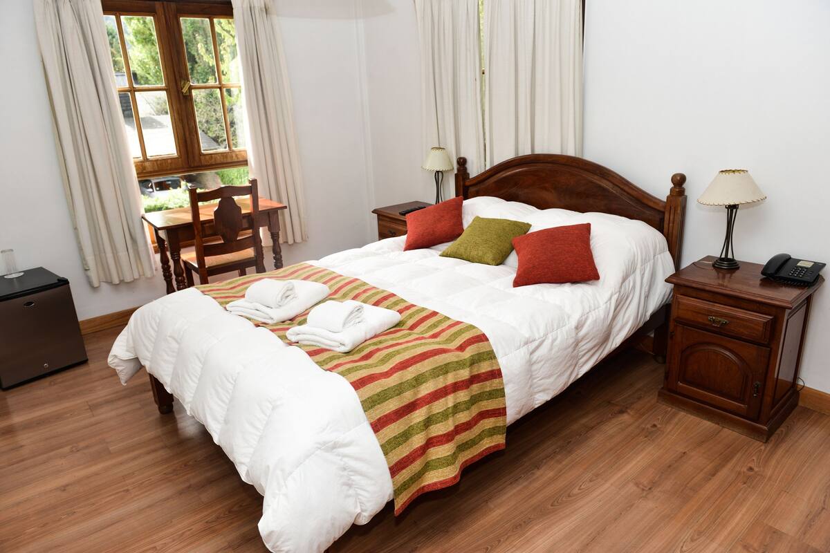 Een net opgemaakt bed met een gestreept dekbed, twee bedlampen, een houten nachtkastje met een telefoon, en een houten bureau met een stoel.