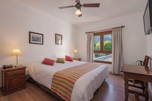 Quarto de hotel com cama, abajures, mesa de cabeceira de madeira, escrivaninha e janela com cortinas.