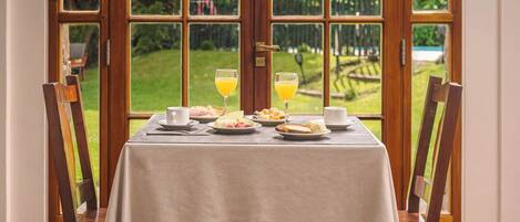 Una mesa de comedor con mantel blanco, dos sillas y vistas a un jardín a través de un ventanal arqueado.