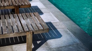 Des fauteuils de jardin en bois sont disposés près d’une piscine, le long d’un chemin bordé de carreaux.