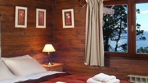 Une chambre à coucher dans une cabane en bois, avec une couvre-lit rouge, des oreillers blancs, une table de chevet avec une lampe et une fenêtre donnant sur des arbres et des montagnes.