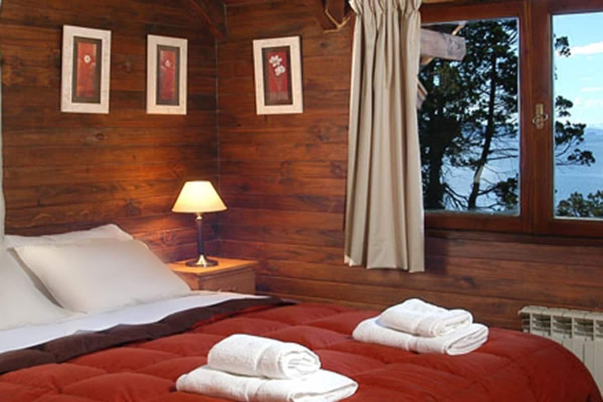 Un dormitorio tipo cabaña de madera, con colcha roja, almohadas blancas, una mesita de noche con lámpara y ventana con vistas a árboles y montañas.