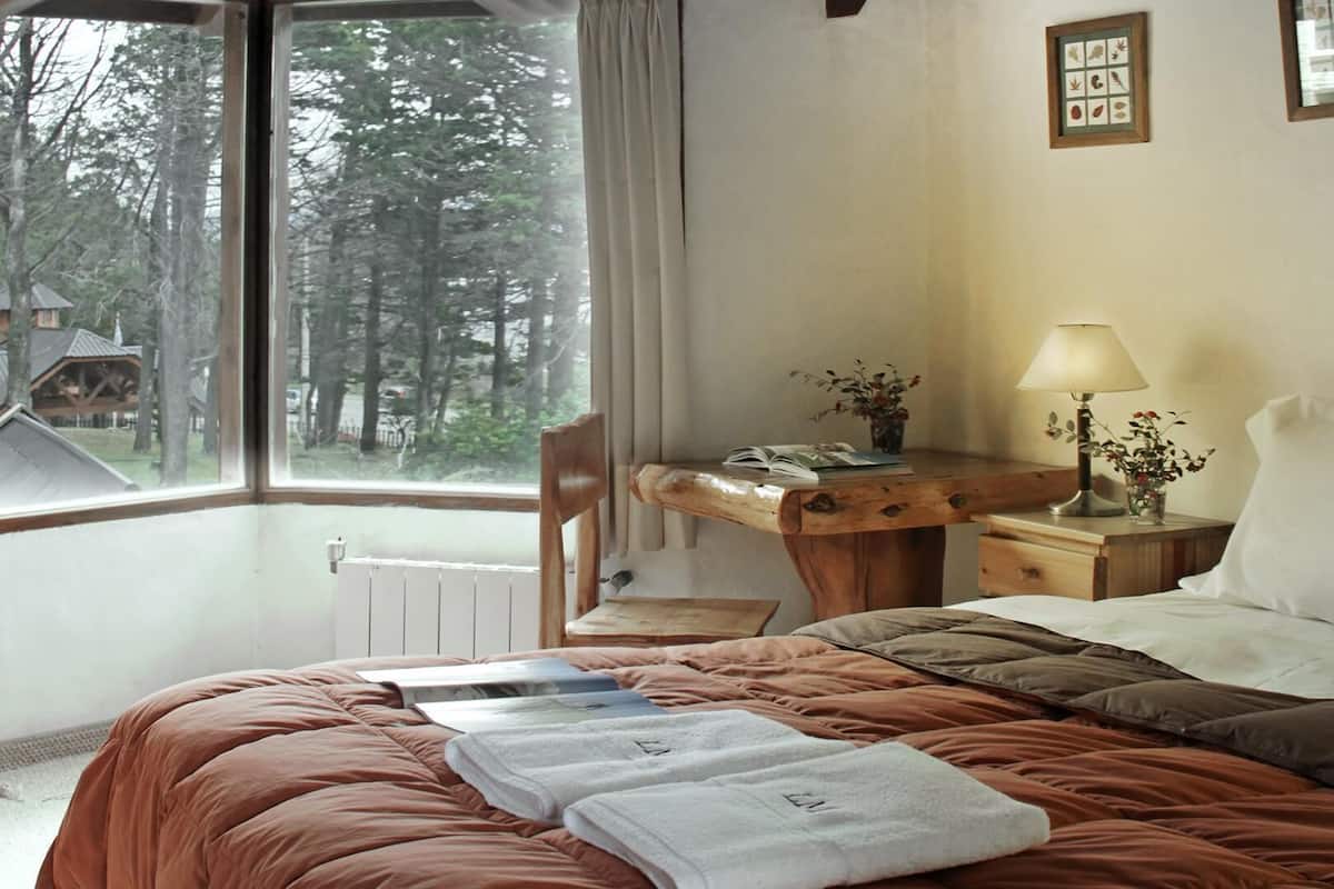 Habitación de hotel con cama, escritorio, silla, mesita de noche, lámpara y ventana con vista a árboles.