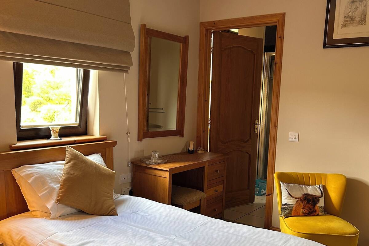 Quarto com cama, cômoda de madeira, cadeira amarela, espelho e janela com vista para o verde.