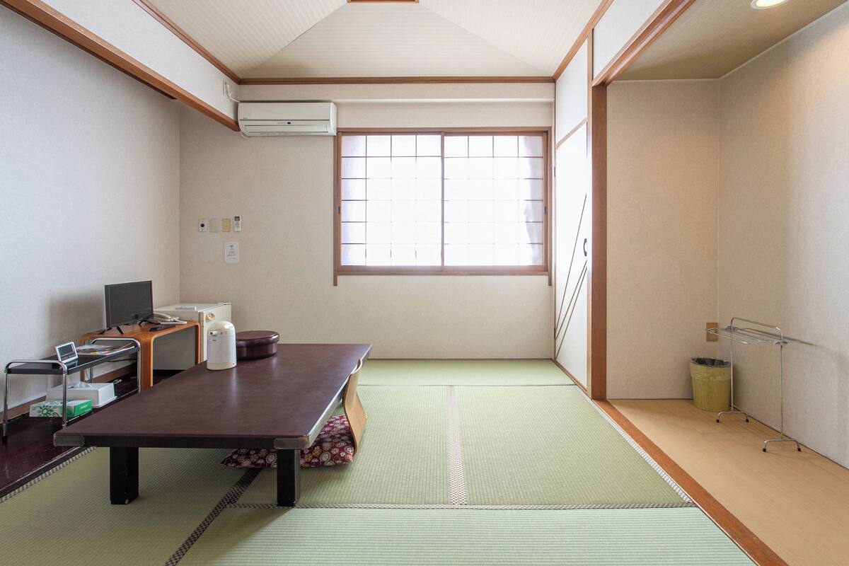 A traditional Japanese room with tatami flooring, a tatami mat table, a low wooden desk with a computer, a window with shoji screens, and a ceiling with recessed lighting.