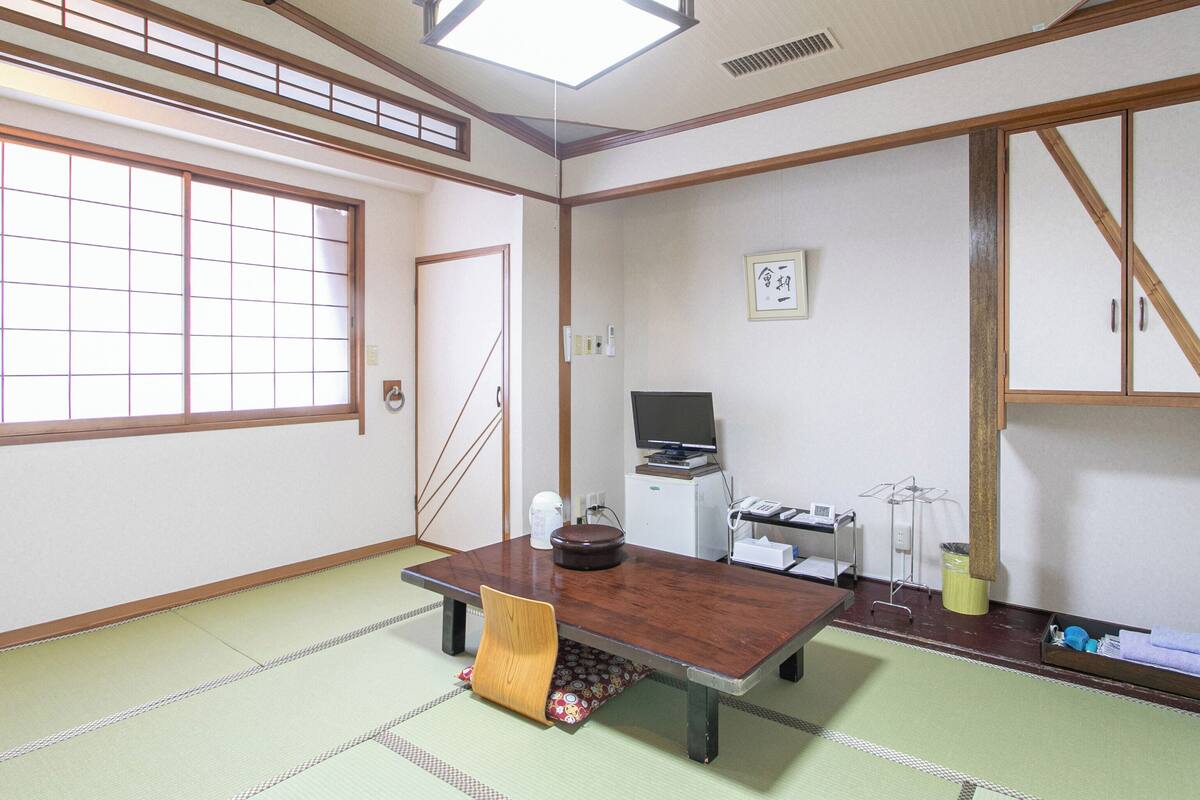 A traditional Japanese room with tatami flooring, a low table with a cushion, a television on a stand, and a shoji screen door.