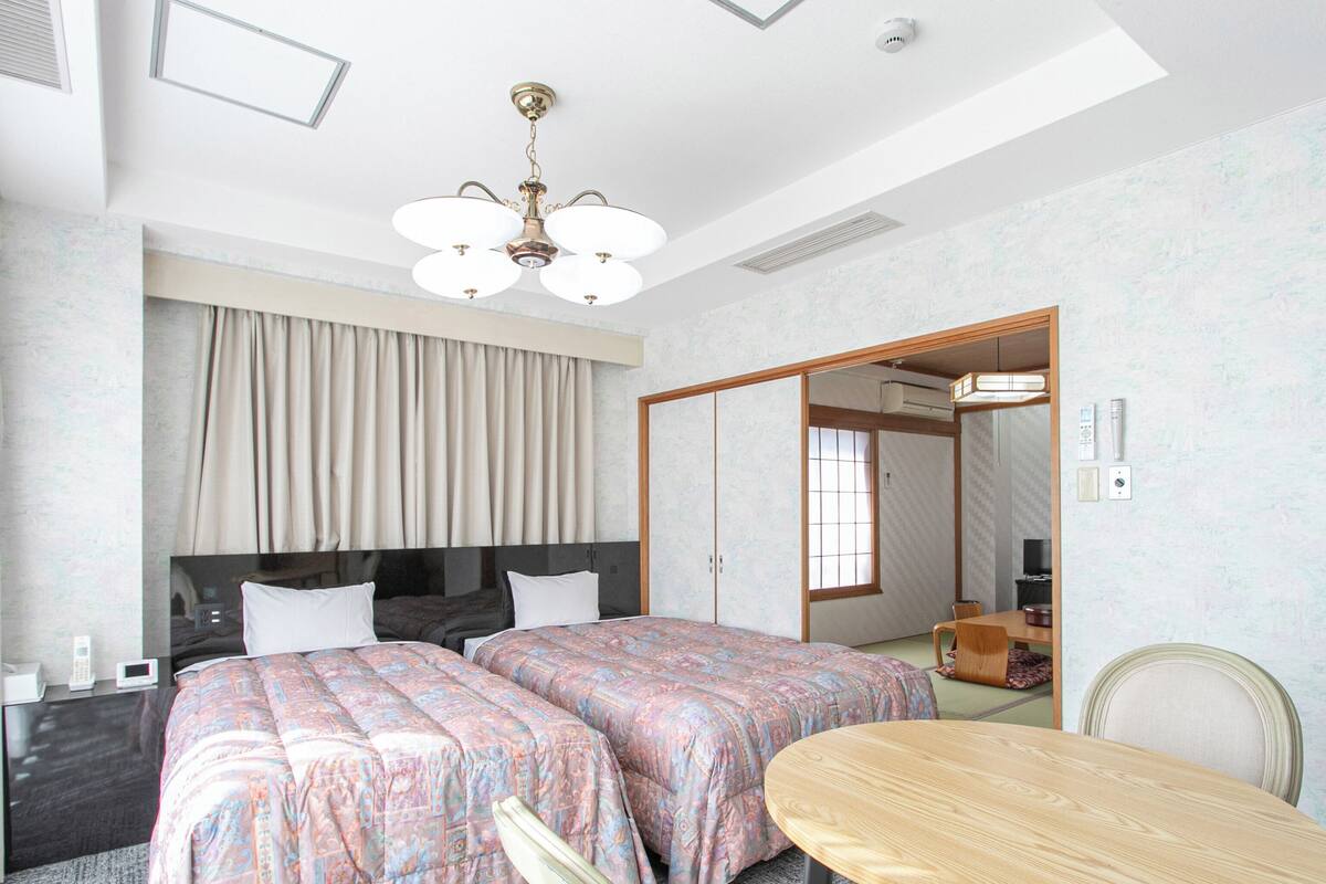 A hotel room with two beds, a desk, a chair, a table, a ceiling fan, and a sliding door.