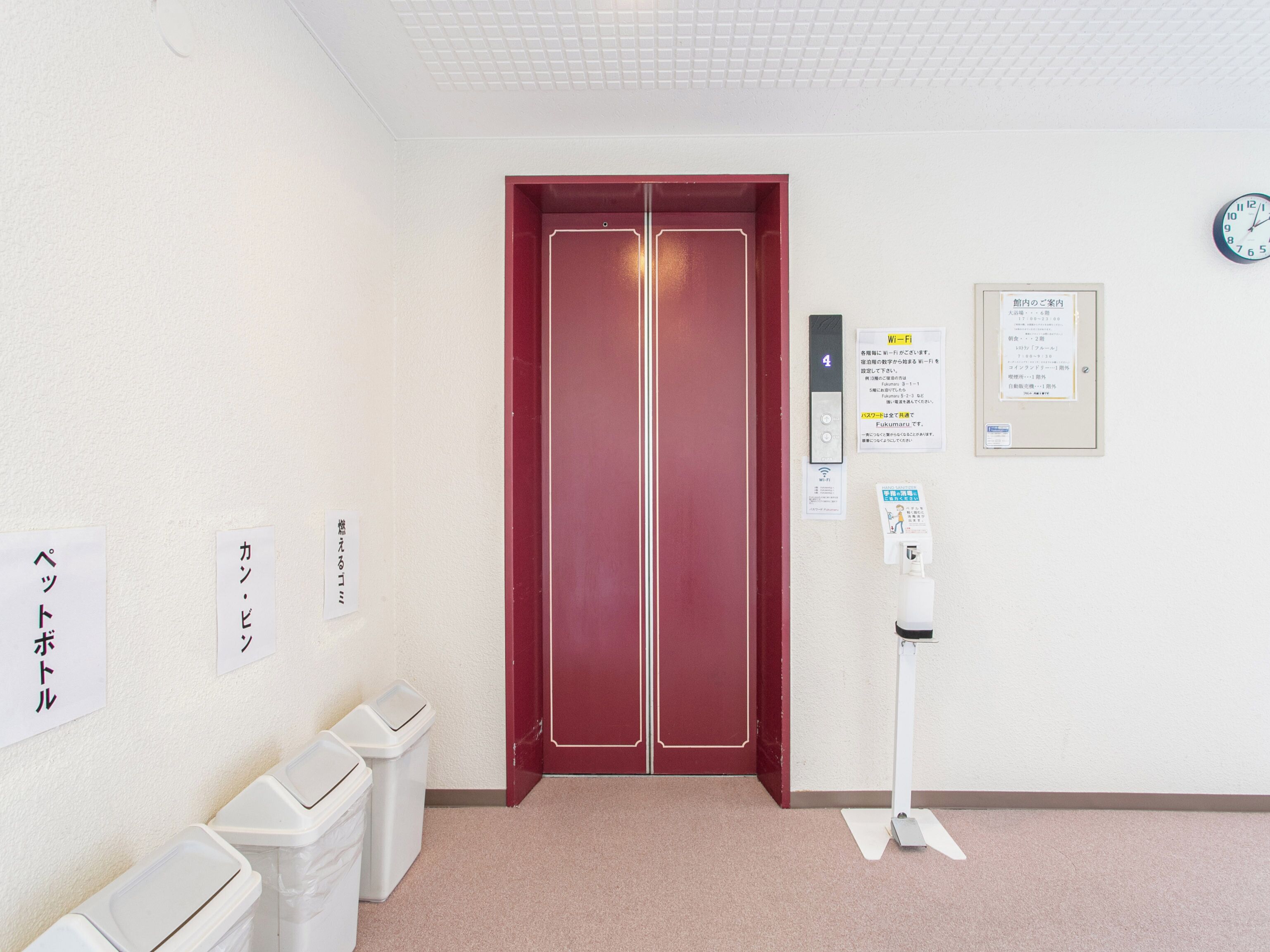 A hotel entrance with a red door, trash bins, a Wi-Fi sign, a clock, and a notice board.