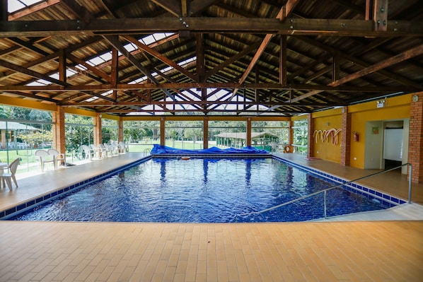 Una piscina interior con techo de madera, suelo de baldosas y vistas a un área verde exterior.