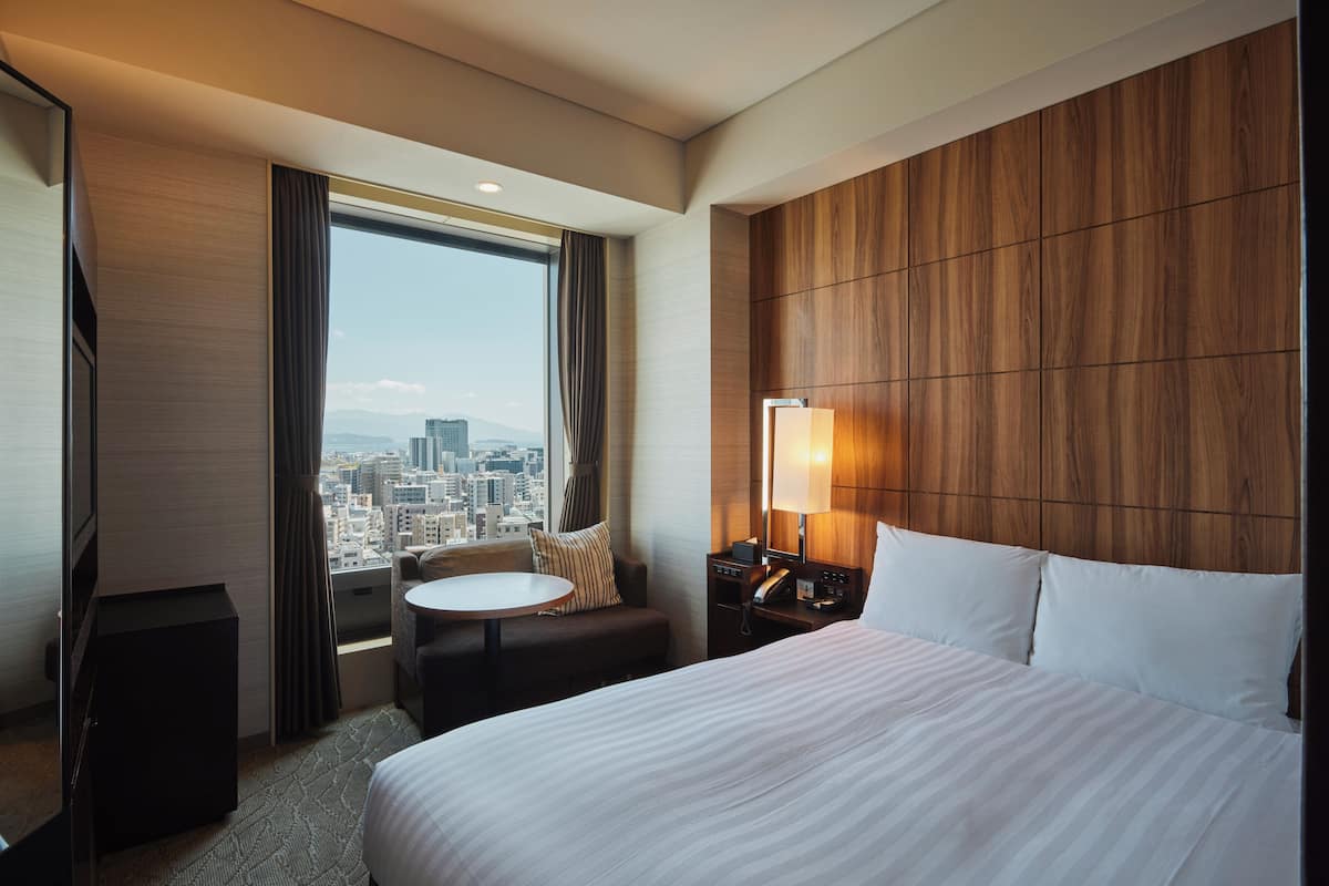 A hotel room with a large bed, a desk with a lamp, a small round table, and a view of the city through a large window.