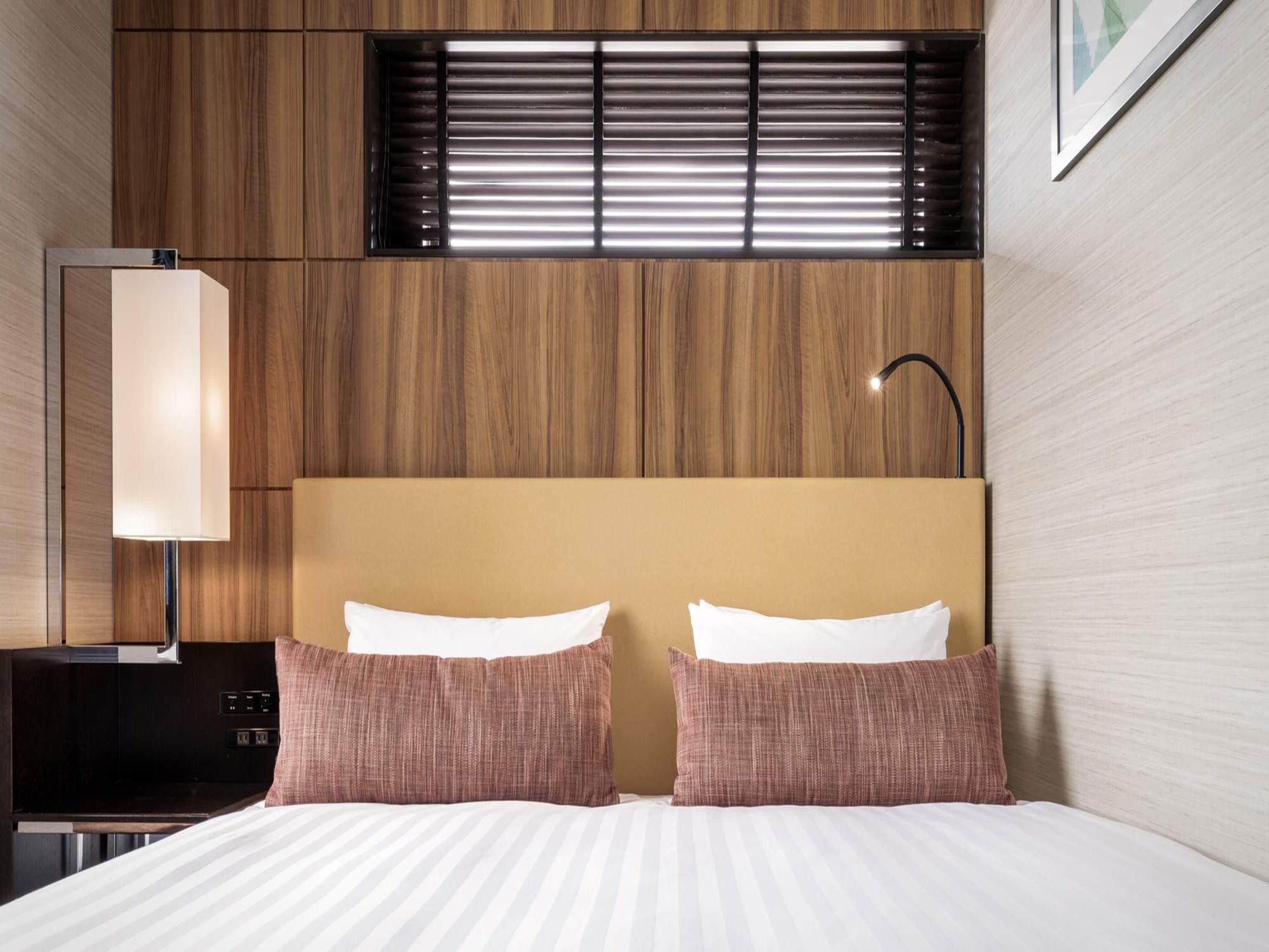 A hotel room with a bed, two pillows, a nightstand, a lamp, and a window with blinds.