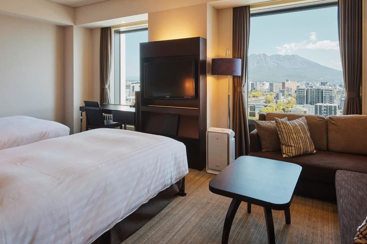 A hotel room with two beds, a sofa, a TV, and a view of a cityscape with mountains.