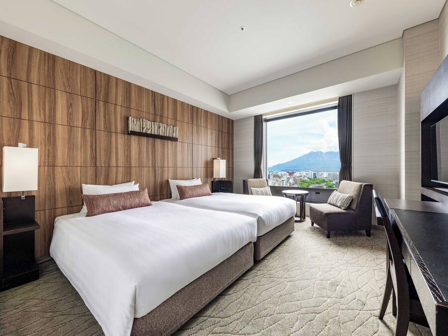 Een hotelkamer met twee bedden, een bureau, een stoel en een groot raam met uitzicht op de stad.