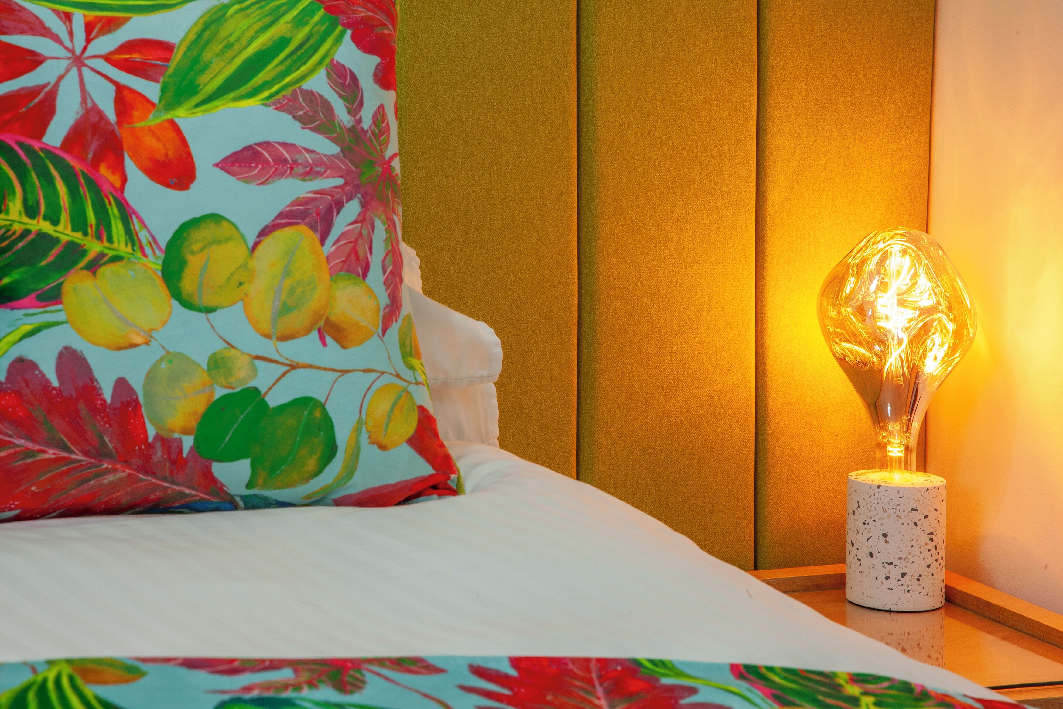 A bed with a colorful floral pillow, a textured wall, and a decorative light fixture.