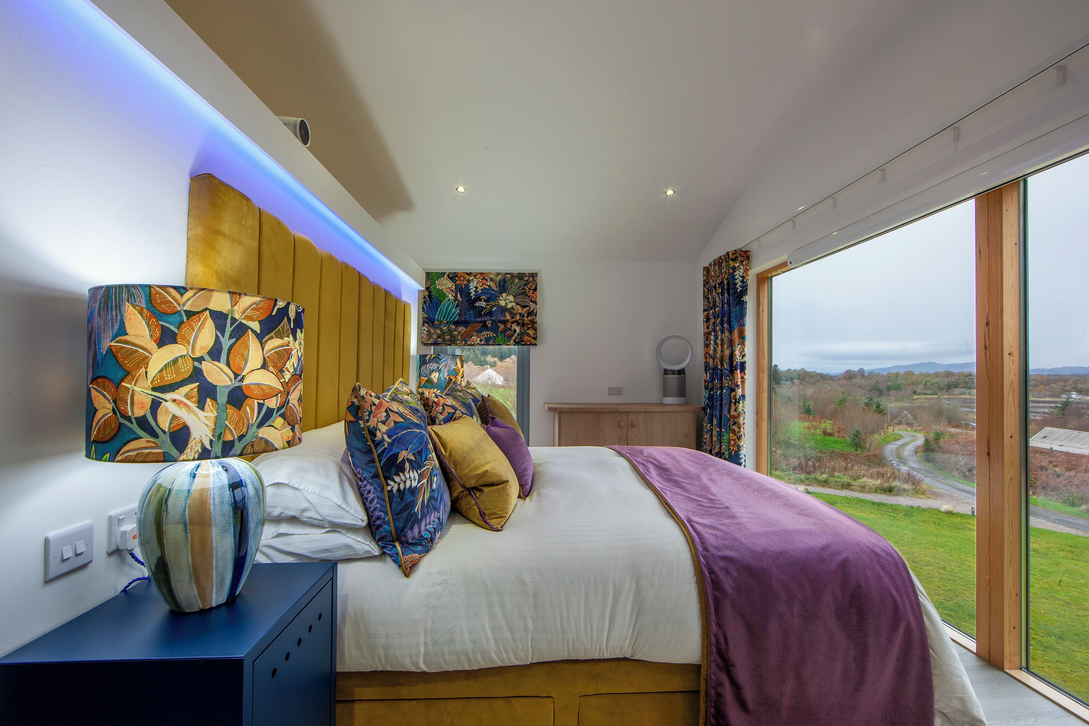A modern bedroom with a large bed, colorful pillows, a vase, and a view of the countryside through large windows.