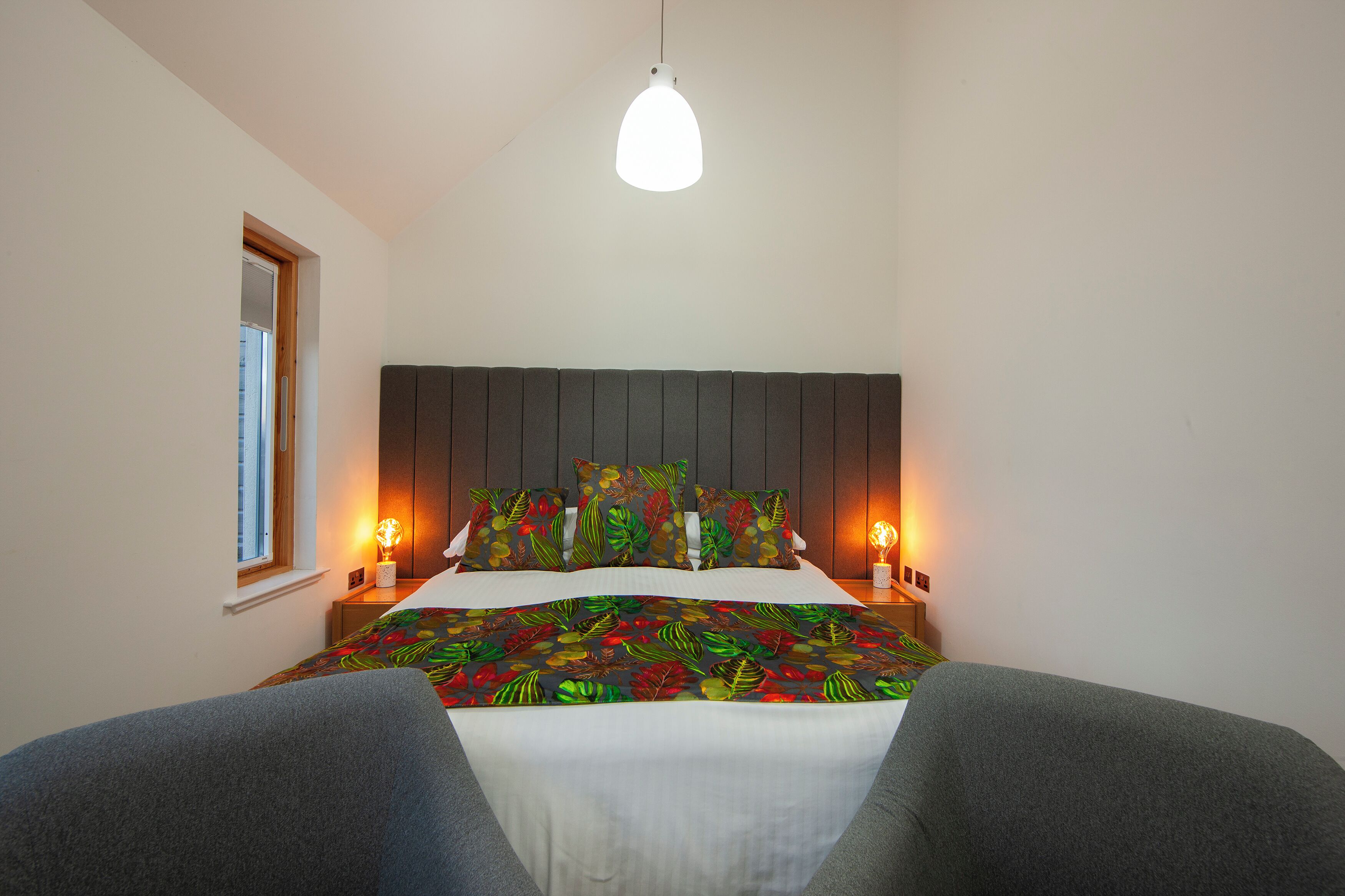 Un dormitorio con cama, dos mesitas de noche, una ventana y una lámpara colgante.