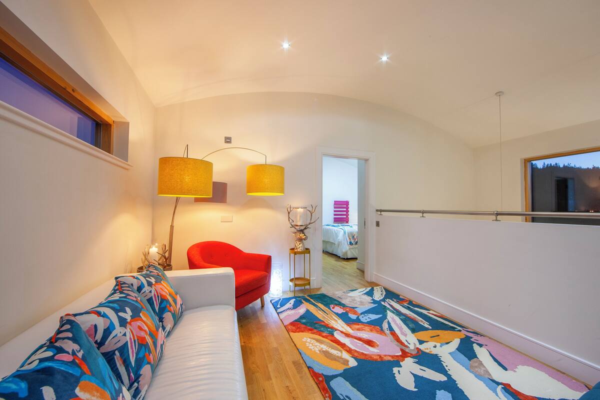 A modern living room with a white sofa, a red chair, and a colorful rug. There is a small table with a lamp and a decorative item. The room has a wooden floor and a large window with a view of the outside.