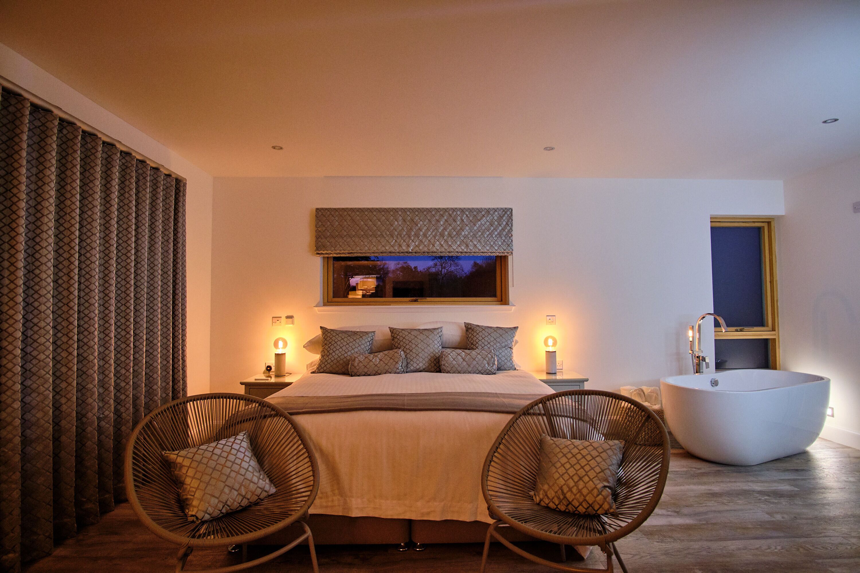 A bedroom with a large bed, two wicker chairs, a built-in bathtub, and a large window with a view.